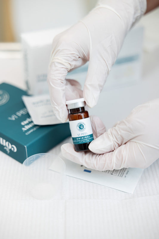 Gloved hands holding a vial of VI Peel Original chemical peel solution, with accompanying skincare materials and packaging visible in the background on a clinical treatment tray.