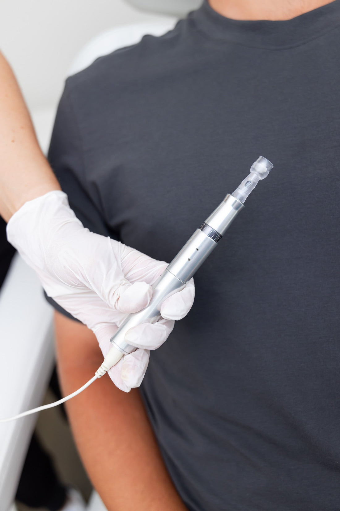A close-up of a gloved hand holding a microneedling device next to a patient in a dark shirt, preparing for a skin treatment.