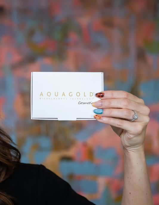 A hand with colorful, artistic nails holding a white AQUAGOLD microchannel technology cosmetic box against a colorful abstract background.