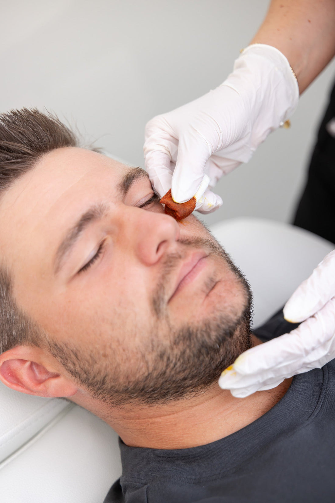 A man receiving a facial treatment while lying down with his eyes closed. A gloved professional is gently applying a treatment or serum under his eye using a cotton pad. The atmosphere appears clean and clinical.