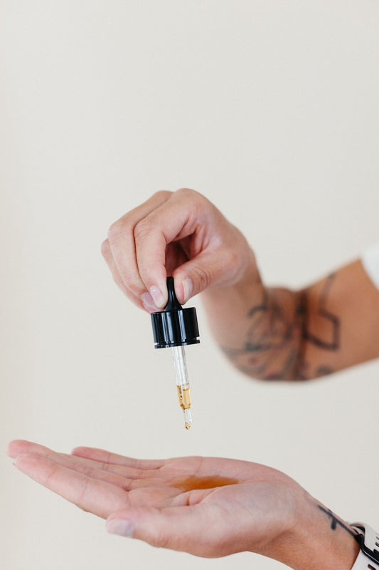 A person with tattooed arms uses a dropper to dispense a golden facial serum into their palm, demonstrating skincare application against a neutral background