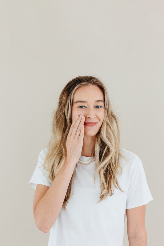 A smiling woman with long blonde hair wearing a white t-shirt gently applies skincare product to her cheek, showcasing a natural and fresh-faced look against a neutral background.