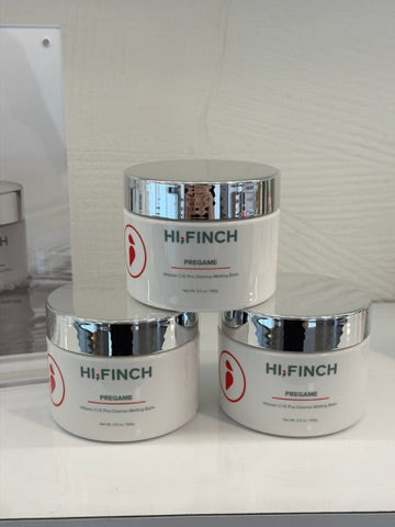 Three white jars of Hi, Finch Pregame Vitamin C+E pre-cleanse melting balm with silver lids, arranged on a glass shelf, one behind two in front.
