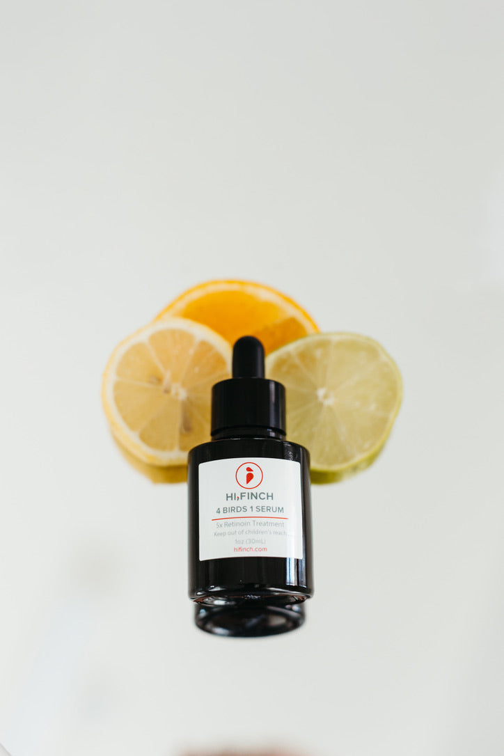 Dark brown serum bottle with black dropper, "H.FINCH 4 BIRDS 1 SERUM," against sliced orange, lemon, and lime on a reflective white surface.