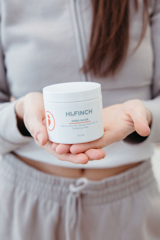 A woman in gray loungewear holds an open palm displaying a white Hi, Finch Speed Racer jar labeled "Glycolic Acid 2% / Salicylic Acid USP, 2% Exfoliating Pads, 60 pads."
