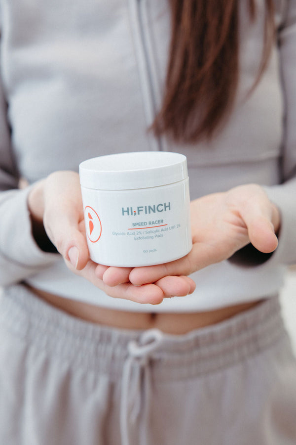 A woman in gray loungewear holds an open palm displaying a white Hi, Finch Speed Racer jar labeled "Glycolic Acid 2% / Salicylic Acid USP, 2% Exfoliating Pads, 60 pads."