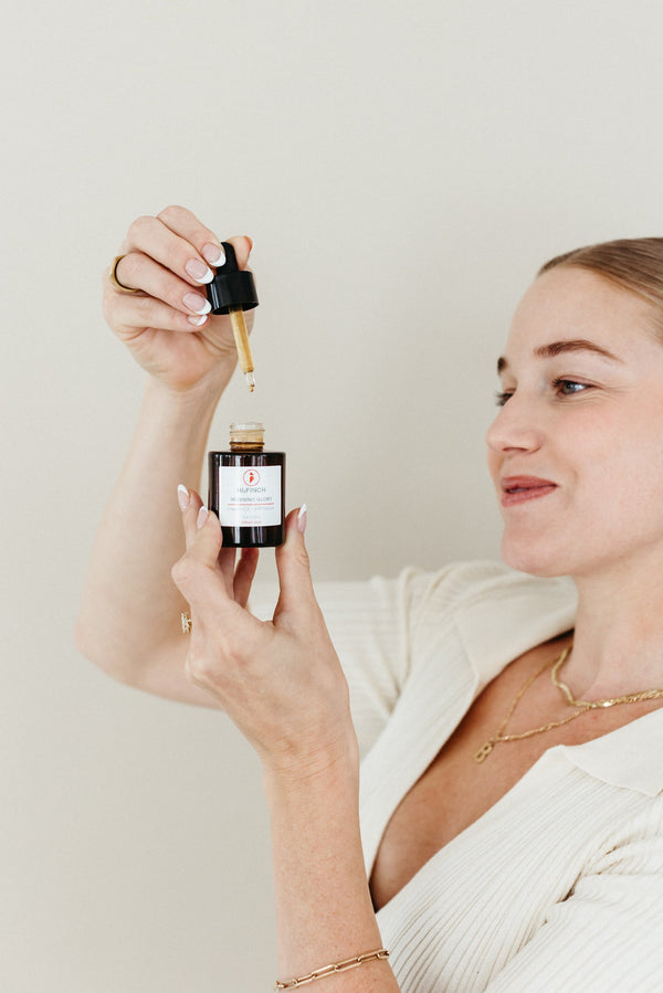 A woman holds a dropper above a dark amber bottle of Hi, Finch Morning Glory vitamin C serum against a neutral white background.