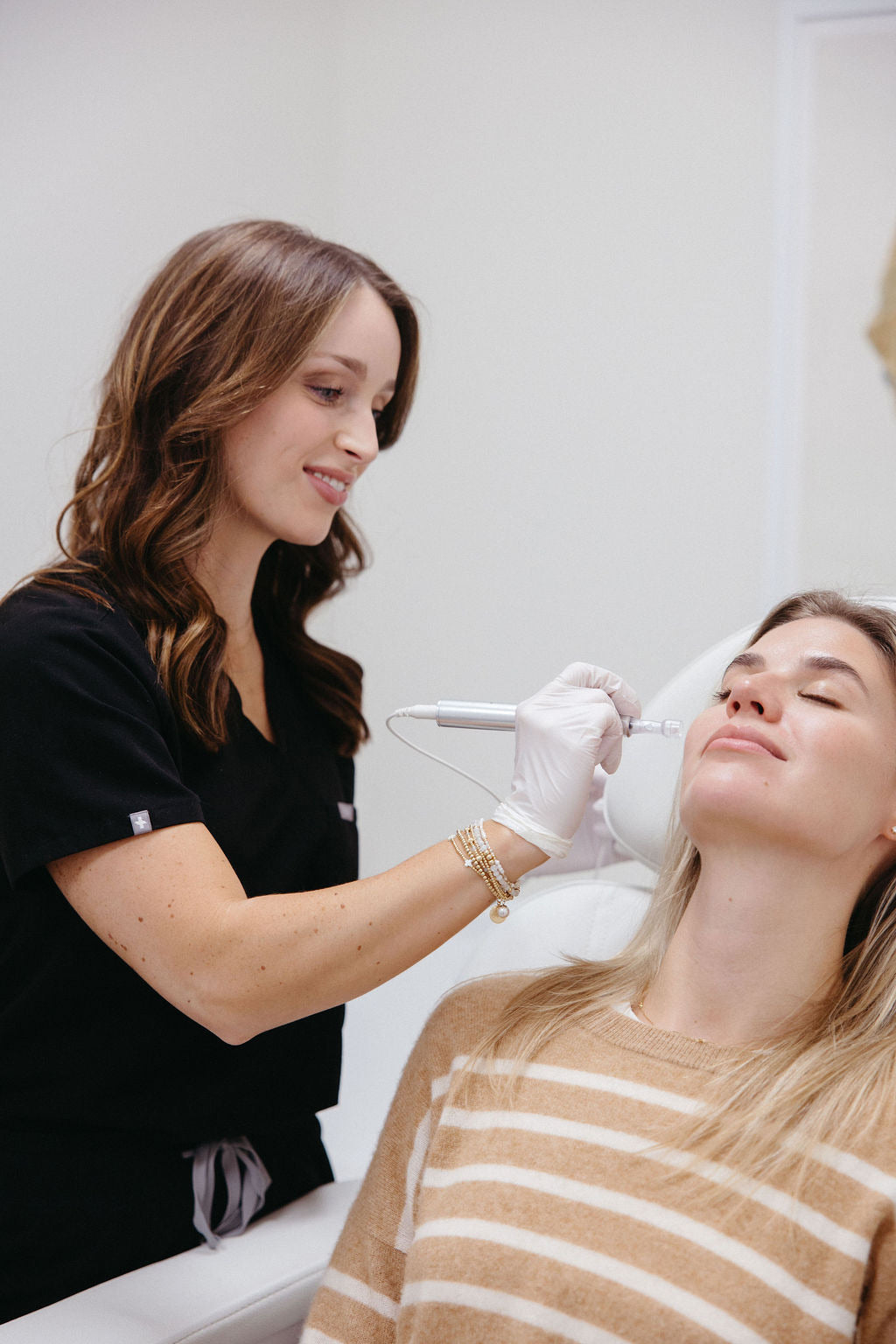 A nurse practitioner in black scrubs performs a microneedling treatment on a smiling patient reclined in a treatment chair.