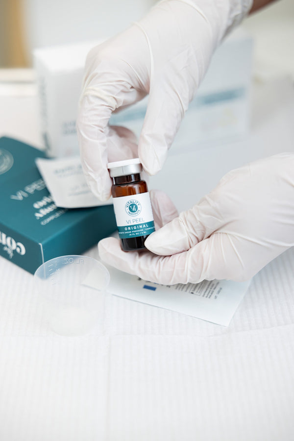A gloved provider holds a small amber vial of a chemical peel along with post procedure kit materials on a treatment surface