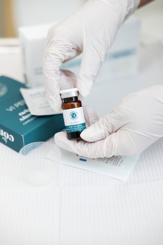 A gloved hand holds a small amber vial labeled "VI Peel Original" over a treatment tray with a measuring cup and Hi, Finch branded boxes in the background.