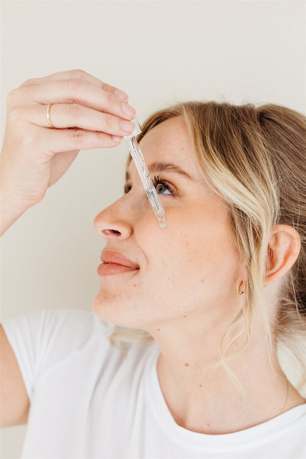A woman with blond hair holds a dropper pipette up to her face while applying a clear serum, photographed in profile against a neutral background.