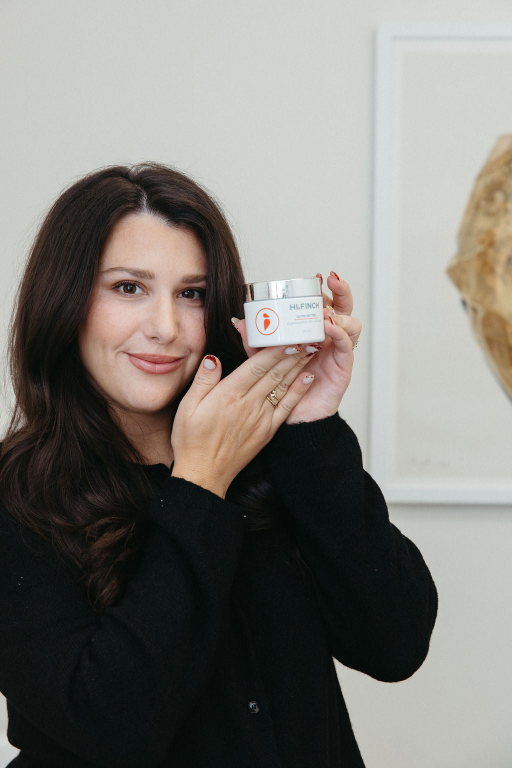 "A woman with long dark hair smiles while holding up a white jar of Hi Finch Glow Getter Brightening Comfort Cream skincare product, wearing a black sweater against a neutral background."