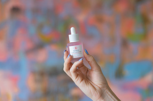 A hand with periwinkle-blue nails holds up a pink glass dropper bottle of Hi, Finch Sunny Disposition Gentle Lactic Acid Resurfacing Serum in front of a blurred, colorful abstract mural.