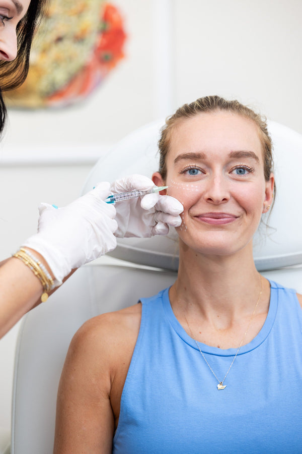 A smiling patient in a treatment chair receives a series of small injections along her cheek from a gloved provider, with numbered injection points marked on her skin.