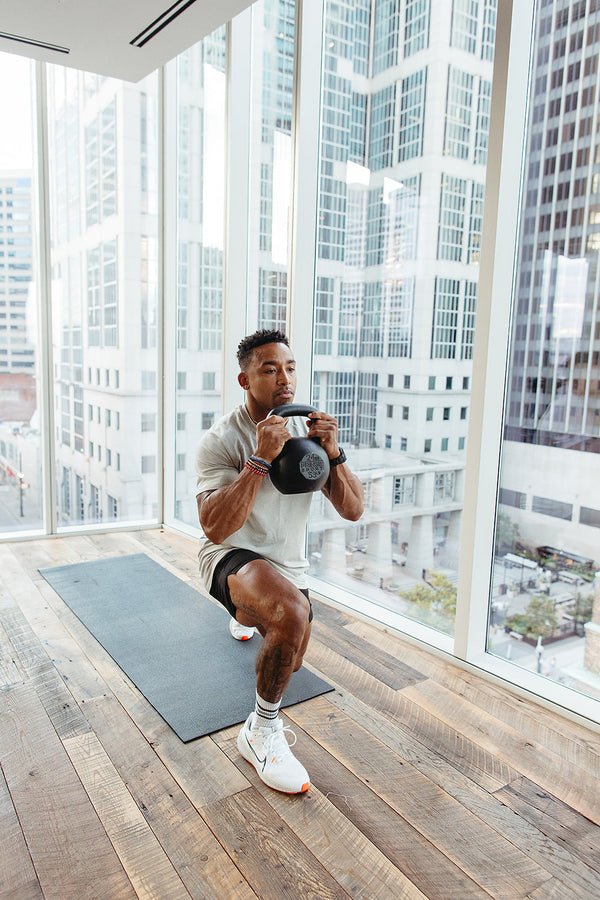 Man doing a kettlebell goblet squat for lower body strength training in a modern gym with floor-to-ceiling windows and urban skyline.