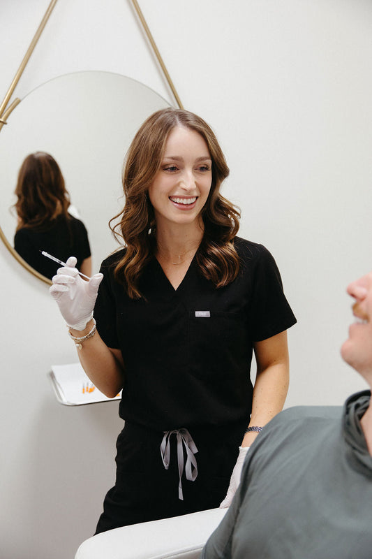A Hi, Finch nurse practitioner in black scrubs smiles while holding a syringe and consulting with a patient in a bright, modern treatment room.