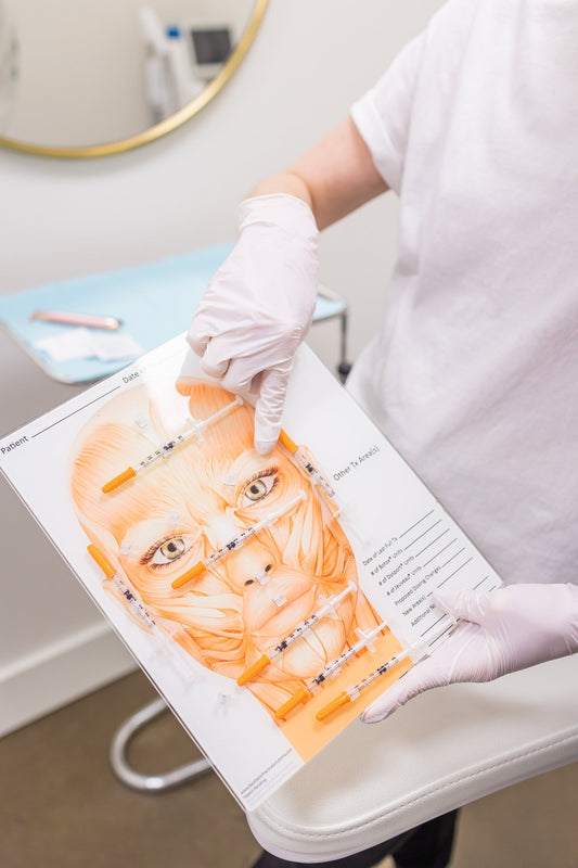 A gloved provider holds a printed facial injection map marked with dosing points, preparing for a Botox treatment in a clinic room.