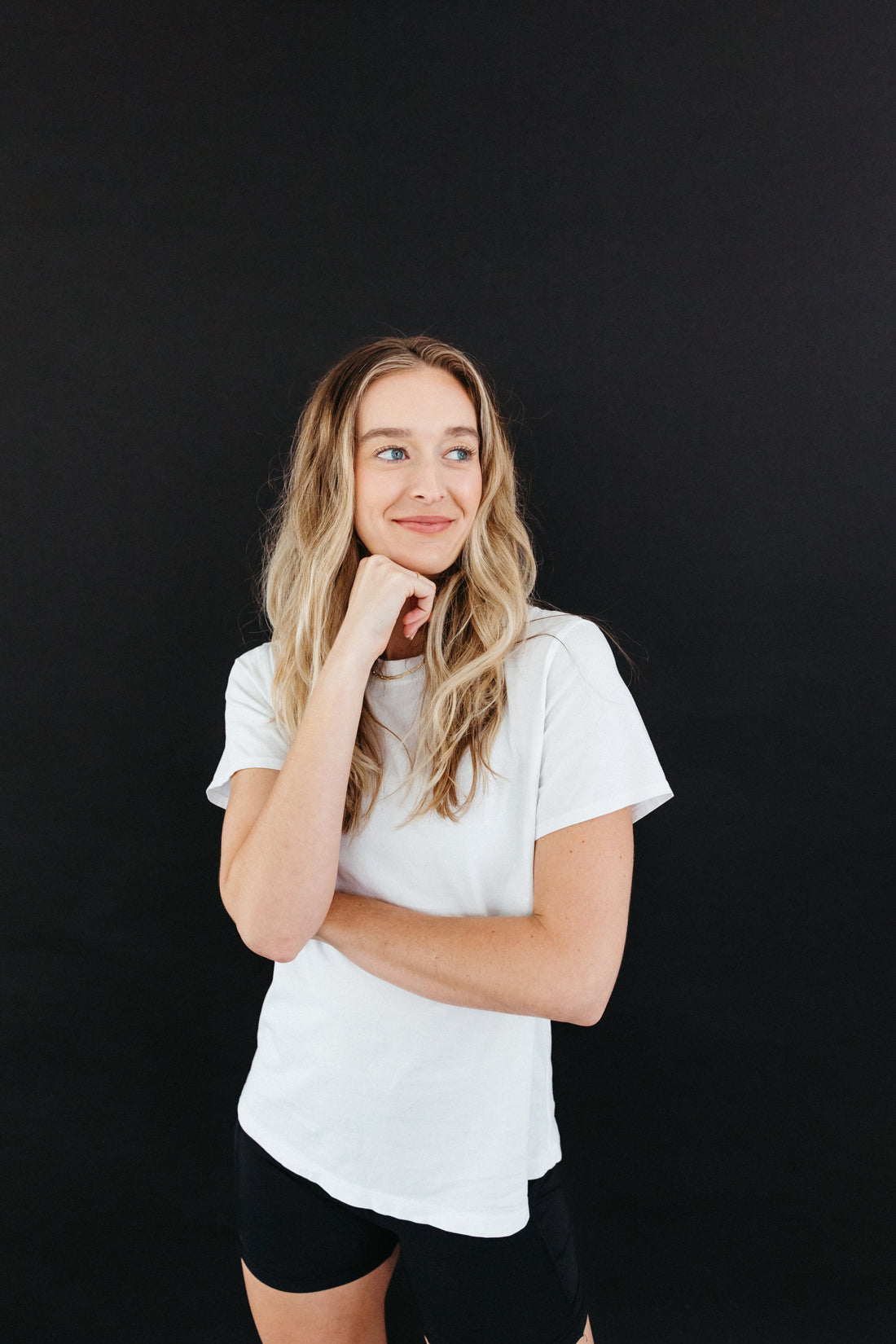 A woman with long, wavy blonde hair wearing a white t-shirt and black shorts poses against a solid black background. She has a thoughtful expression, resting her chin on her hand while smiling slightly.