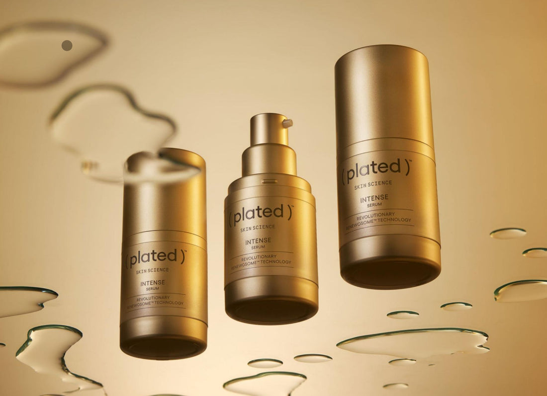 Three gold bottles of Plated Intense Serum are displayed against a golden background with reflective liquid droplets, highlighting the luxurious skincare product.