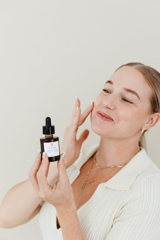 Woman applying facial serum to her cheek while holding a small dropper bottle, smiling against a neutral background.