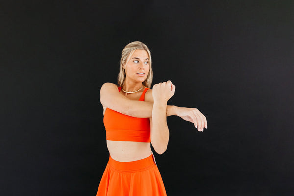 Woman in red bralette and red skirt stretching her arm across her chest.