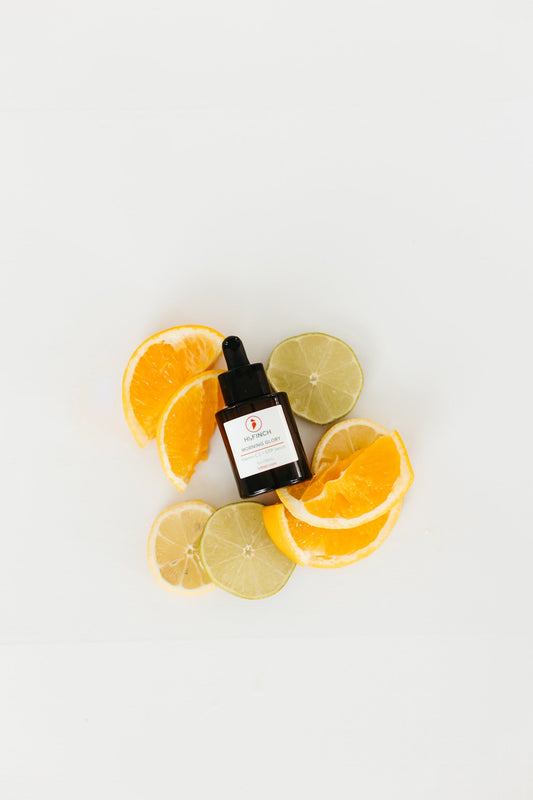 A dark glass dropper bottle of Hi Finch Morning Glory C E + GTP Age Defying Serum placed on a white surface, surrounded by fresh orange, lemon, and lime slices, symbolizing vitamin C–rich ingredients and bright, healthy skin.