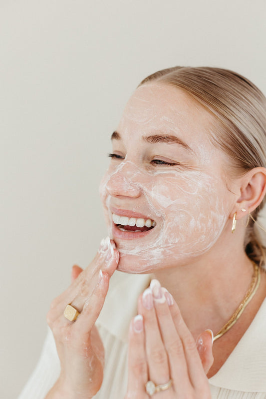 Woman smiling while applying facial cleanser, showing a glowing skincare routine with clean, fresh skin and a minimalist gold jewelry look.