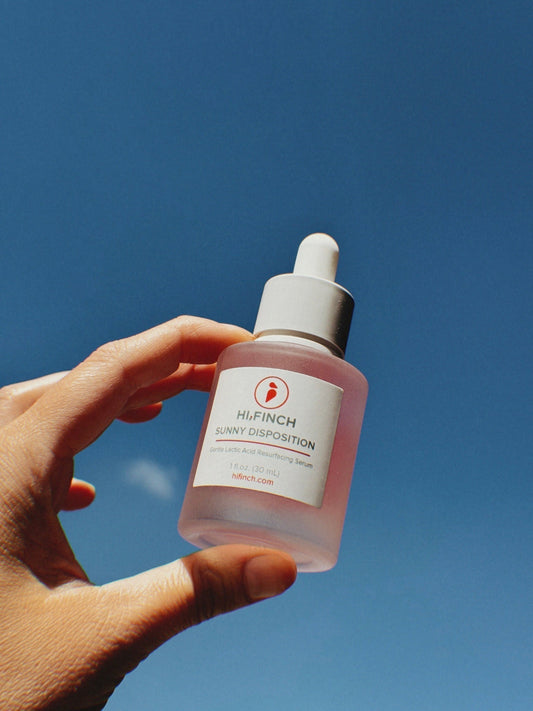 A hand holding a bottle of Hi, Finch Sunny Disposition Gentle Lactic Acid Resurfacing Serum against a bright blue sky. The bottle is frosted with a white dropper cap and a clean, minimal label.