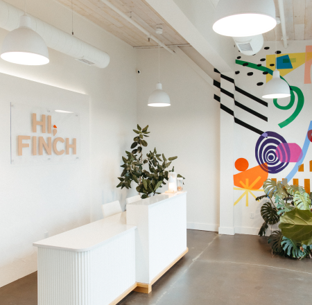 Front desk and reception area at Hi, Finch Cosmetic Dermatology in Nashville, featuring a modern, light-filled interior with white minimalist counters, greenery, and a colorful abstract mural.