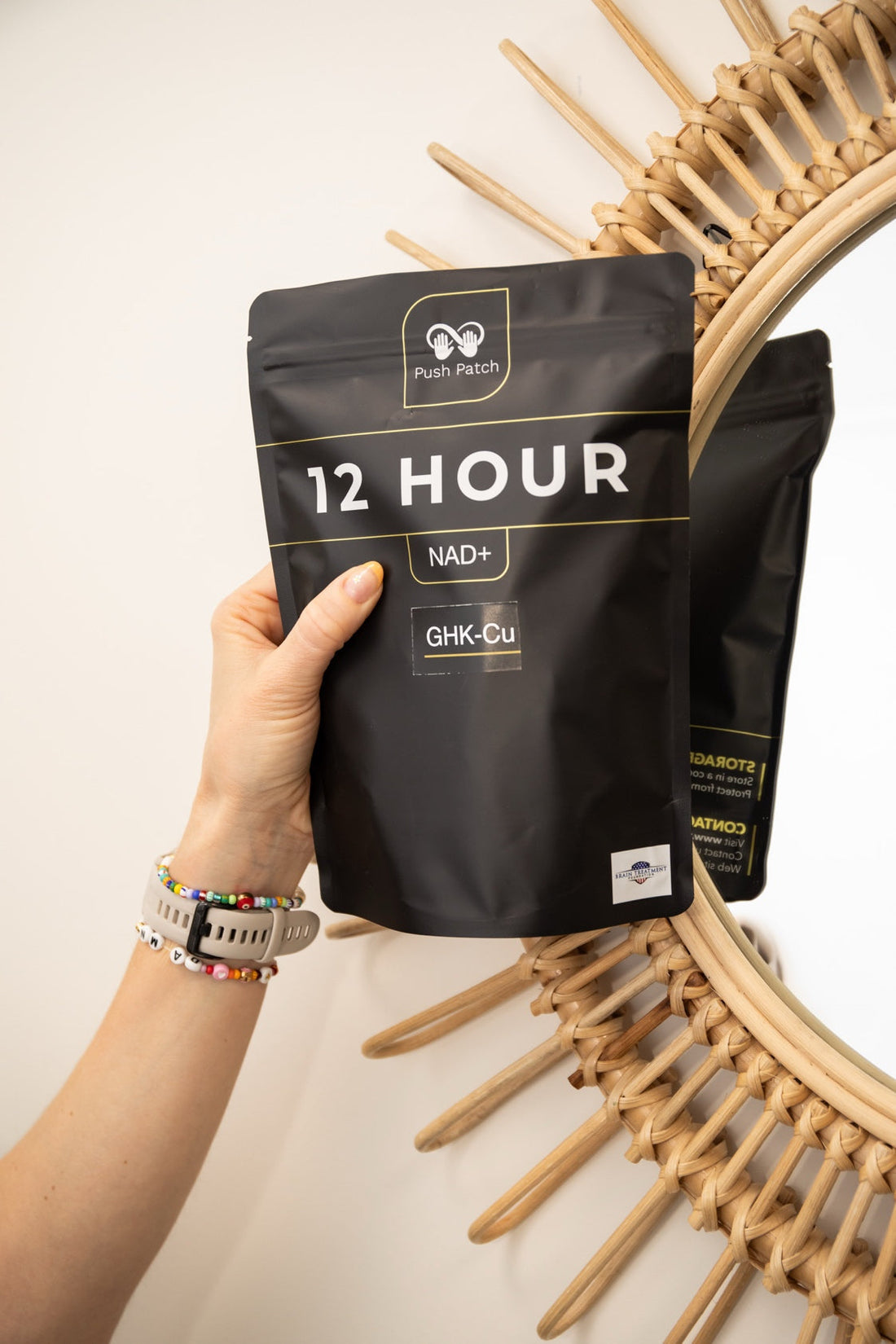 Hand holding a black Push Patch 12 Hour NAD+ and GHK-Cu peptide patch package in front of a round woven rattan mirror, with a second package partially visible in the background.