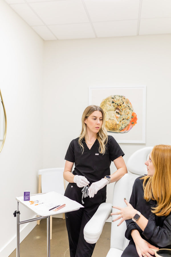  A nurse practitioner in black scrubs and gloves consults with a patient seated in a treatment chair, discussing aesthetic treatment options in a modern, well-lit clinic setting.