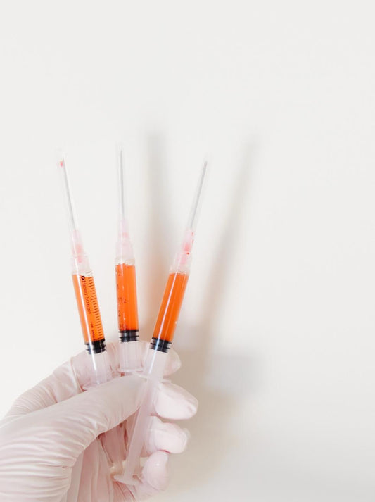 A gloved hand holding three syringes filled with orange platelet-rich fibrin (PRF) solution, prepared for under-eye or facial rejuvenation treatments, against a clean white background.
